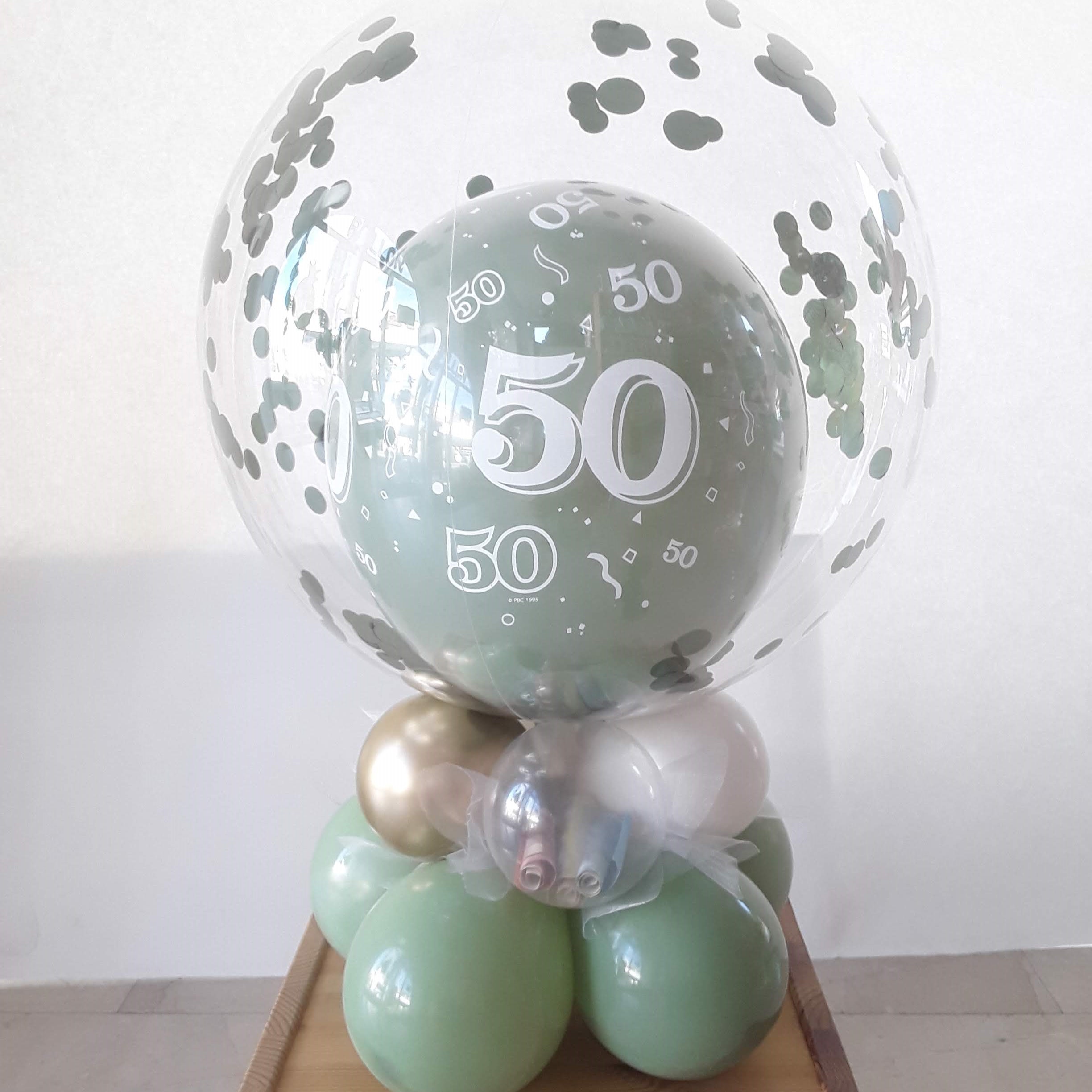 Ballondeko mit durchsichtiger Kugel und grünem 50er-Ballon, gefüllt mit grünem Konfetti, auf Basis aus grünen und goldenen Ballons.