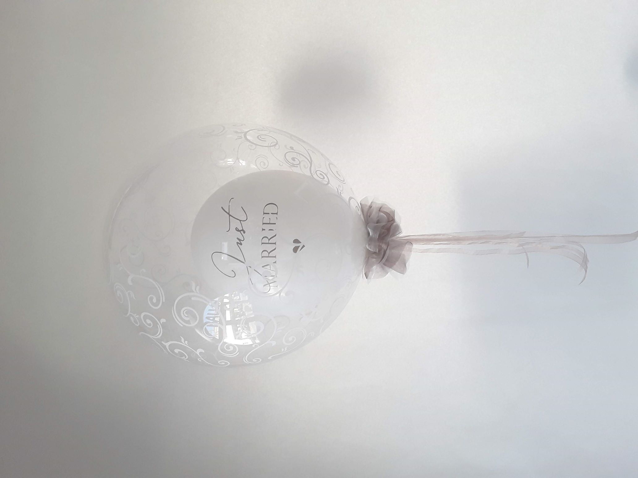 Transparenter Bubble-Ballon mit weißem Innenballon und der Aufschrift „Just Married“, verziert mit weißen Ornamenten und einer eleganten Schleife aus Tüll.