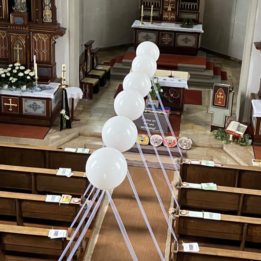 Reihe schwebender weißer Heliumballons mit Bändern, die beidseitig des roten Teppichs in einer Kirche angeordnet sind.-4
