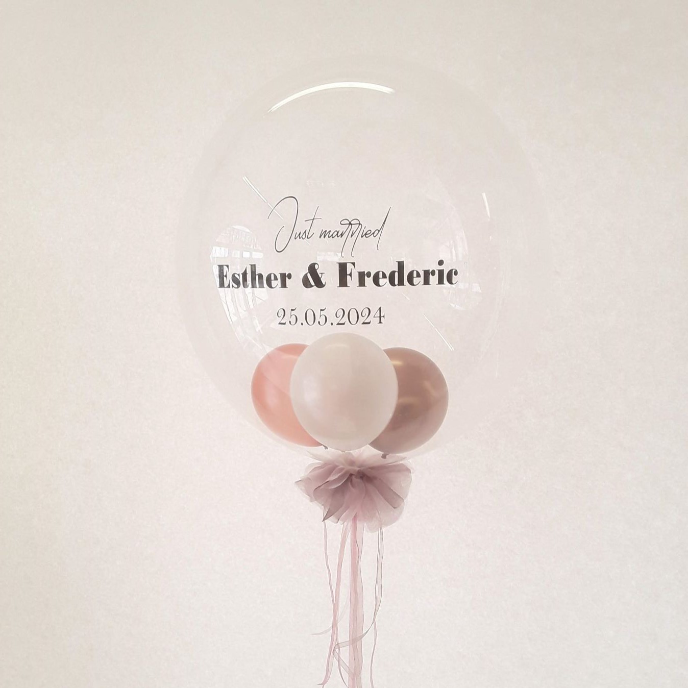 Klarer Ballon mit personalisiertem Aufdruck „Just married Esther & Frederic 25.05.2024“ und drei schwebenden Ballons in Rosa, Weiß und Kupfer sowie einer Schleife aus Tüll.