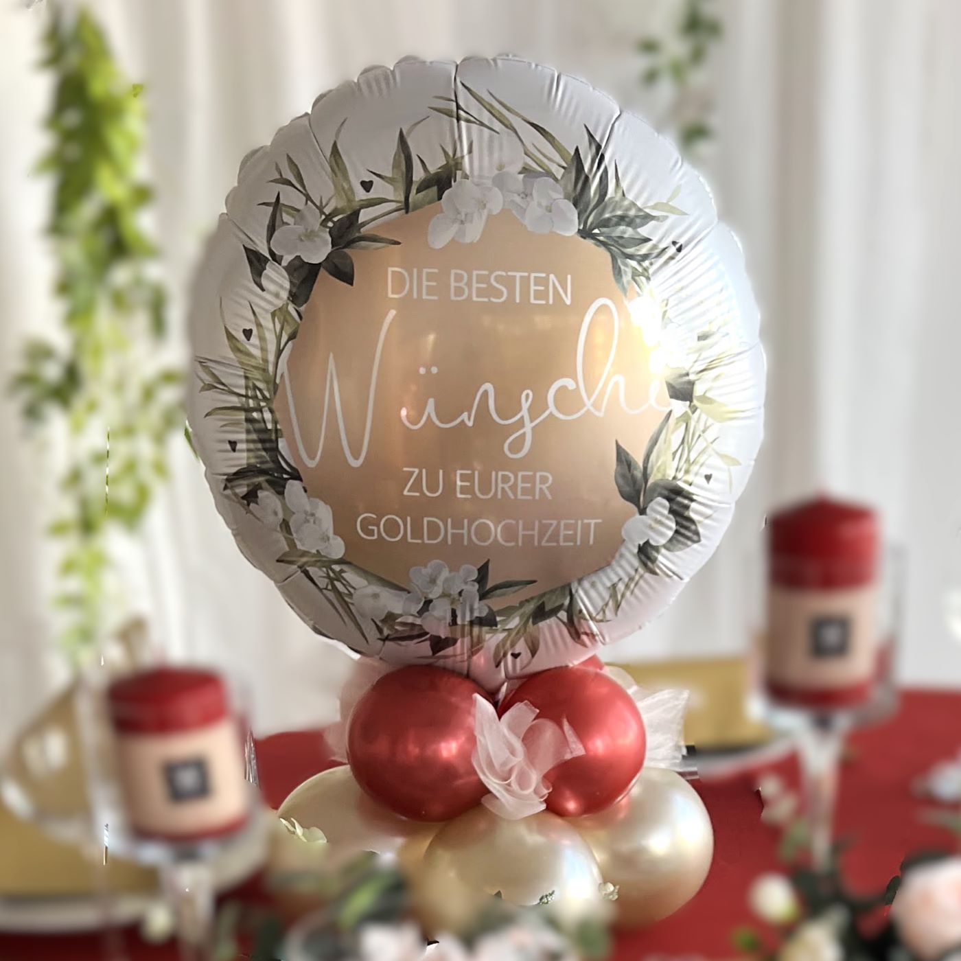 Runder Folienballon in Weiß-Gold mit Blumenkranz und Text 'Die besten Wünsche zu eurer Goldhochzeit', auf rotem und goldenem Ballon-Cluster als Tischbouquet arrangiert.