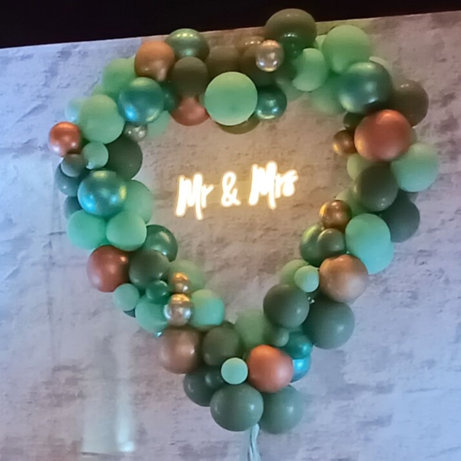 Herzförmiger Ballonbogen in vielen Farben mit unterschiedlich großen Ballons dekoriert und „Mr & Mrs“-Schriftzug in der Mitte grün