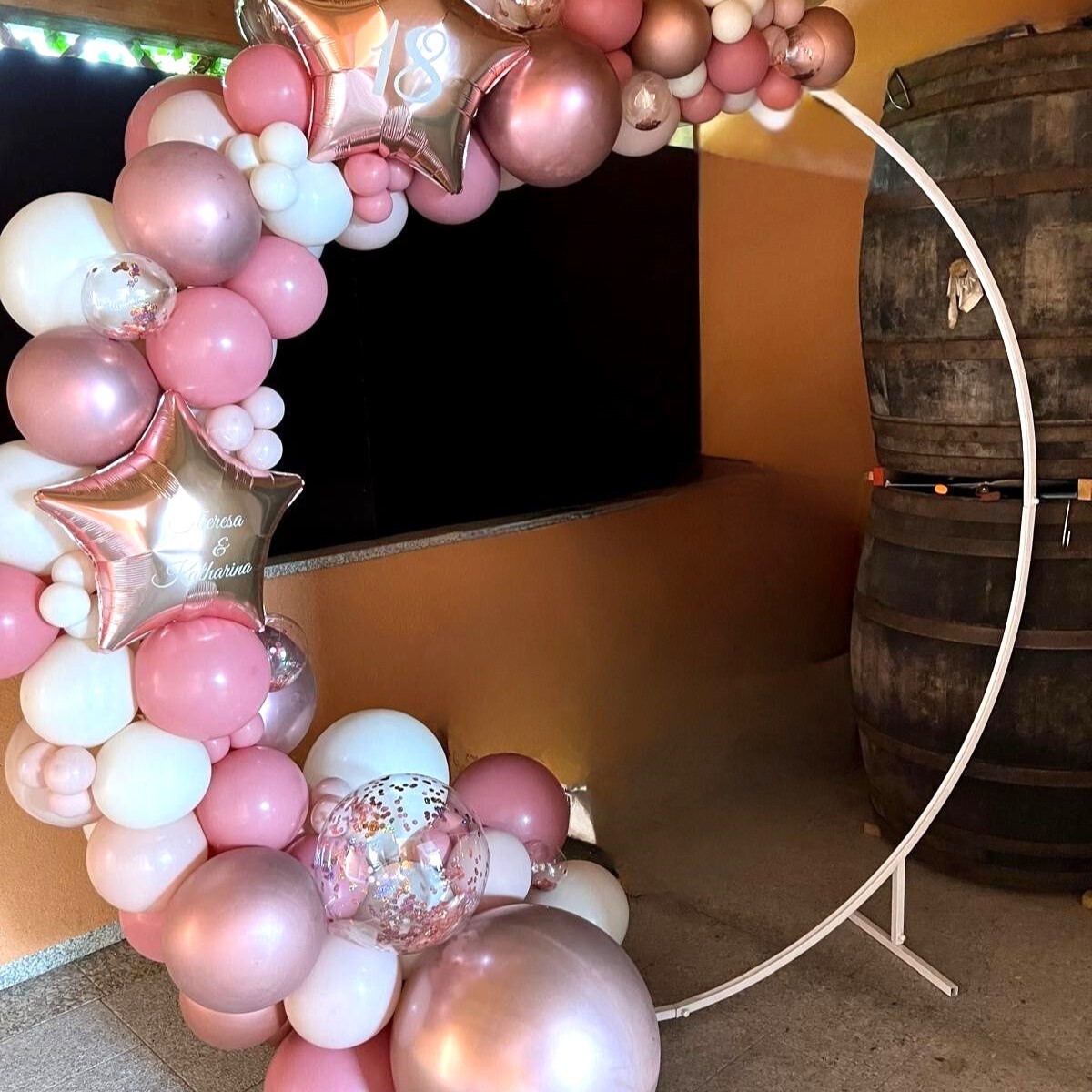 Ringbogen aus Ballons in Roségold, Rosa, Pink und Weiß, dekoriert mit großen Sternfolienballons mit Aufdruck '18' und 'Theresa&Katharina' und transparenten Konfettiballons, aufgebaut in einem Raum mit Holzdecke und zwei Holzfässen im Hintergrund
