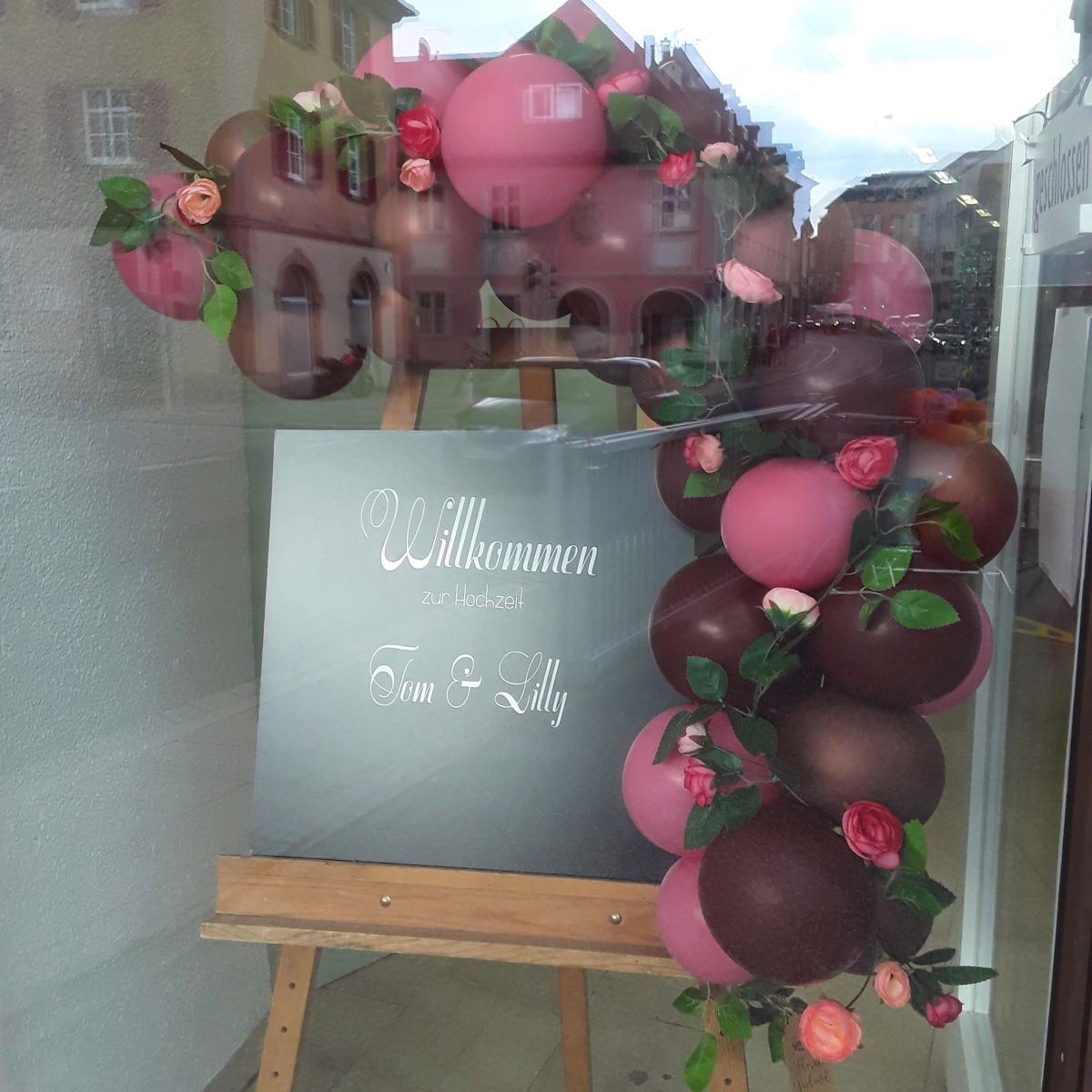 Willkommensschild auf Staffelei mit weißer Aufschrift „Willkommen zur Hochzeit Tom & Lilly“, dekoriert mit Ballongirlande in Rot, Dunkelrot und Rosa sowie künstlichen Rosenblüten und grünen Blättern