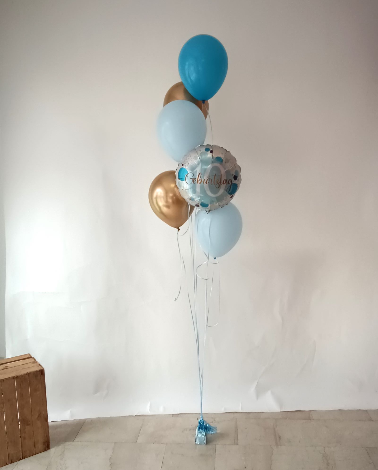 Geburtstags-Ballonbouquet in Blau, Hellblau und Gold mit einem runden silbernen Folienballon mit der Aufschrift 'Geburtstag 10' befestigt an einem Gewicht, Deko-Holzbox