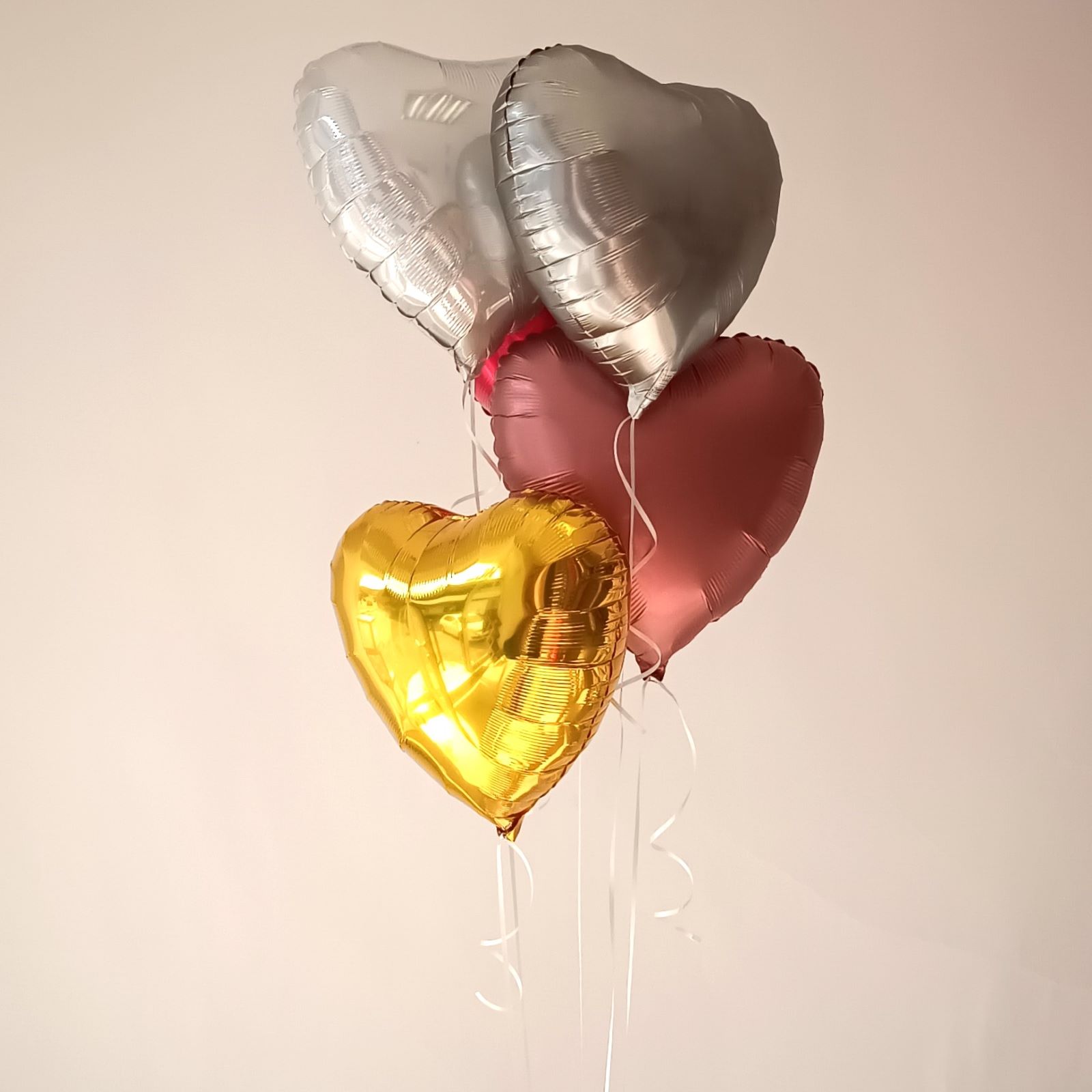 Mehrere Herz-Folienballons in Metallic-Farben (Gold, Silber, Rot, Rosa), mit weißen Bändern, vor neutralem Hintergrund