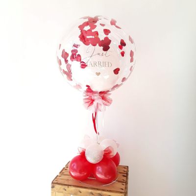 Transparenter Bubble-Ballon mit rotem Herzkonfetti und „Just Married“-Aufdruck, dekoriert mit roter Schleife und auf roter und weißer Ballonbasis mit Glitzereffekt arrangiert.