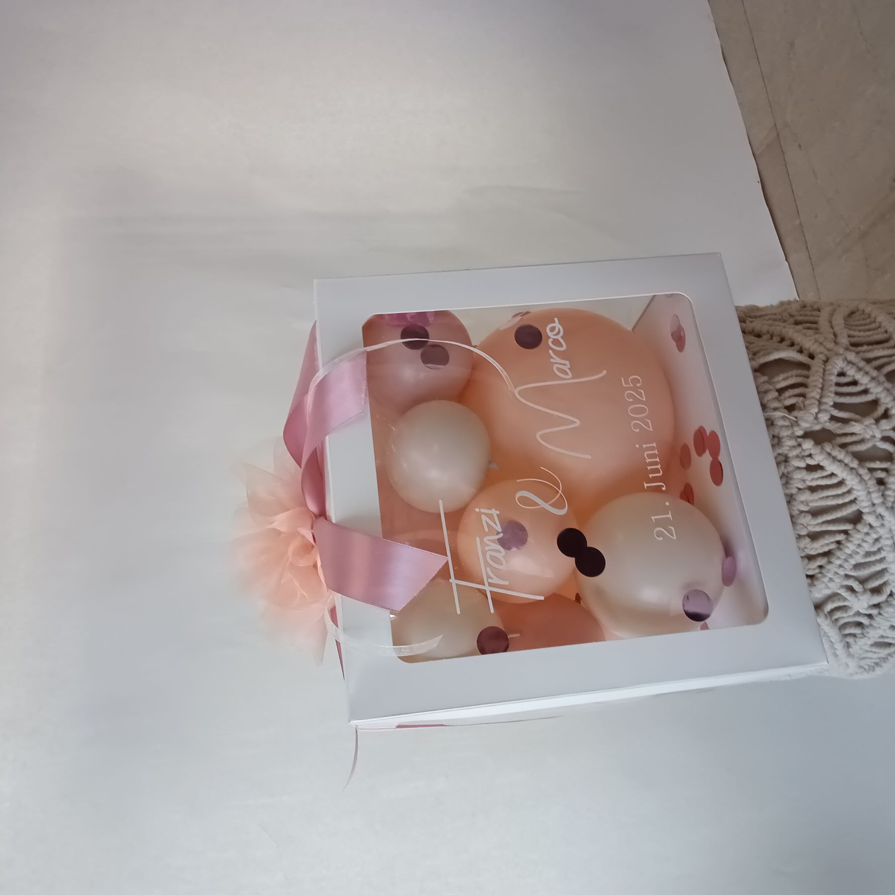 Weiße Ballonbox mit apricot- und roséfarbenen Luftballons, Konfetti, transparenter Front, rosa Satinband und Tüllschleife.