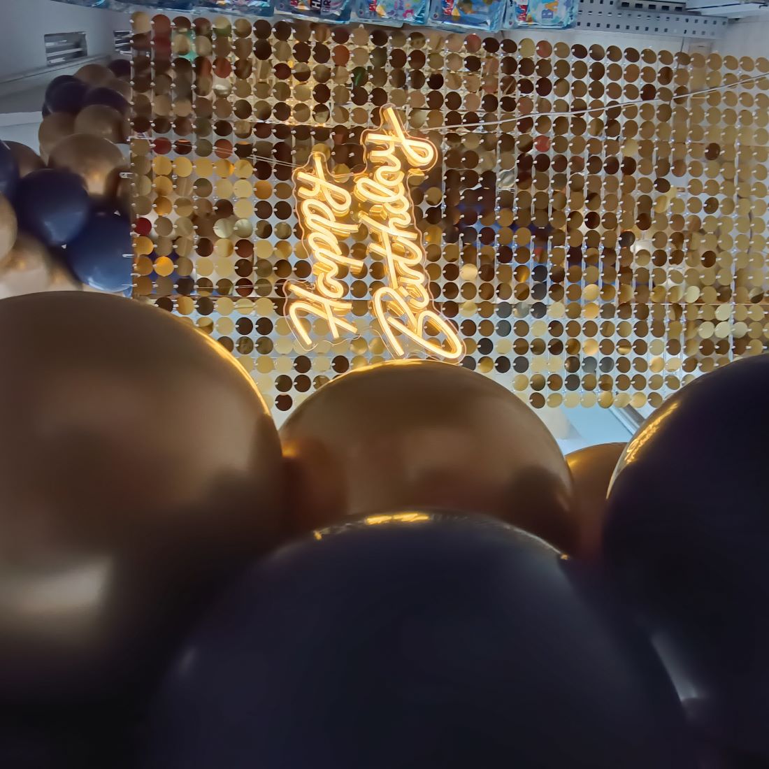 Ausschitt eines Halbbogens aus blauen und goldenen Luftballons vor einem schimmernden Dekovorhang mit Leuchtendem Schriftzug 'Happy Birthday'