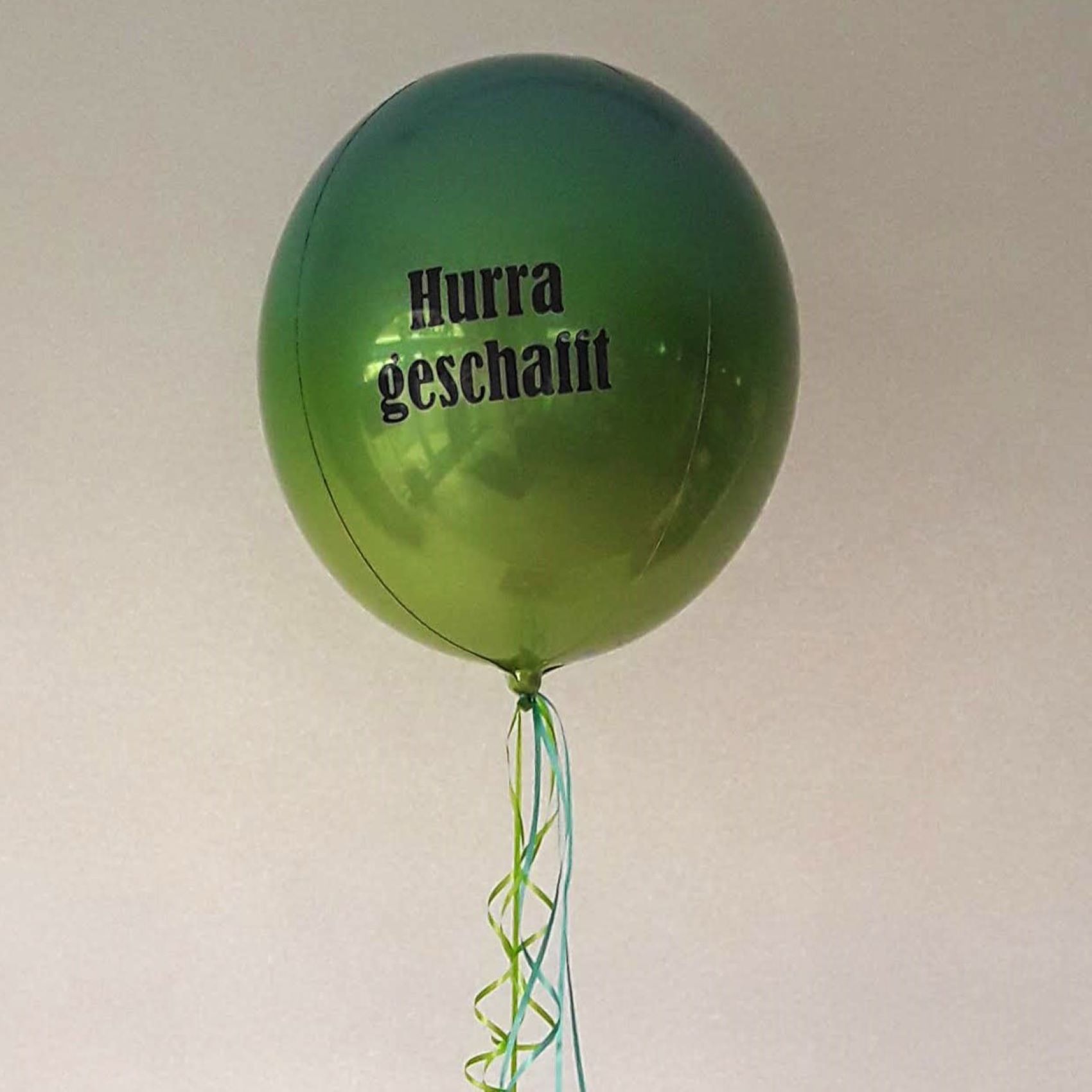Runder, metallisch grüner Orbz-Folienballon mit schwarzem Aufdruck „Hurra geschafft“, daran lange Bänder in Grün und Blau.