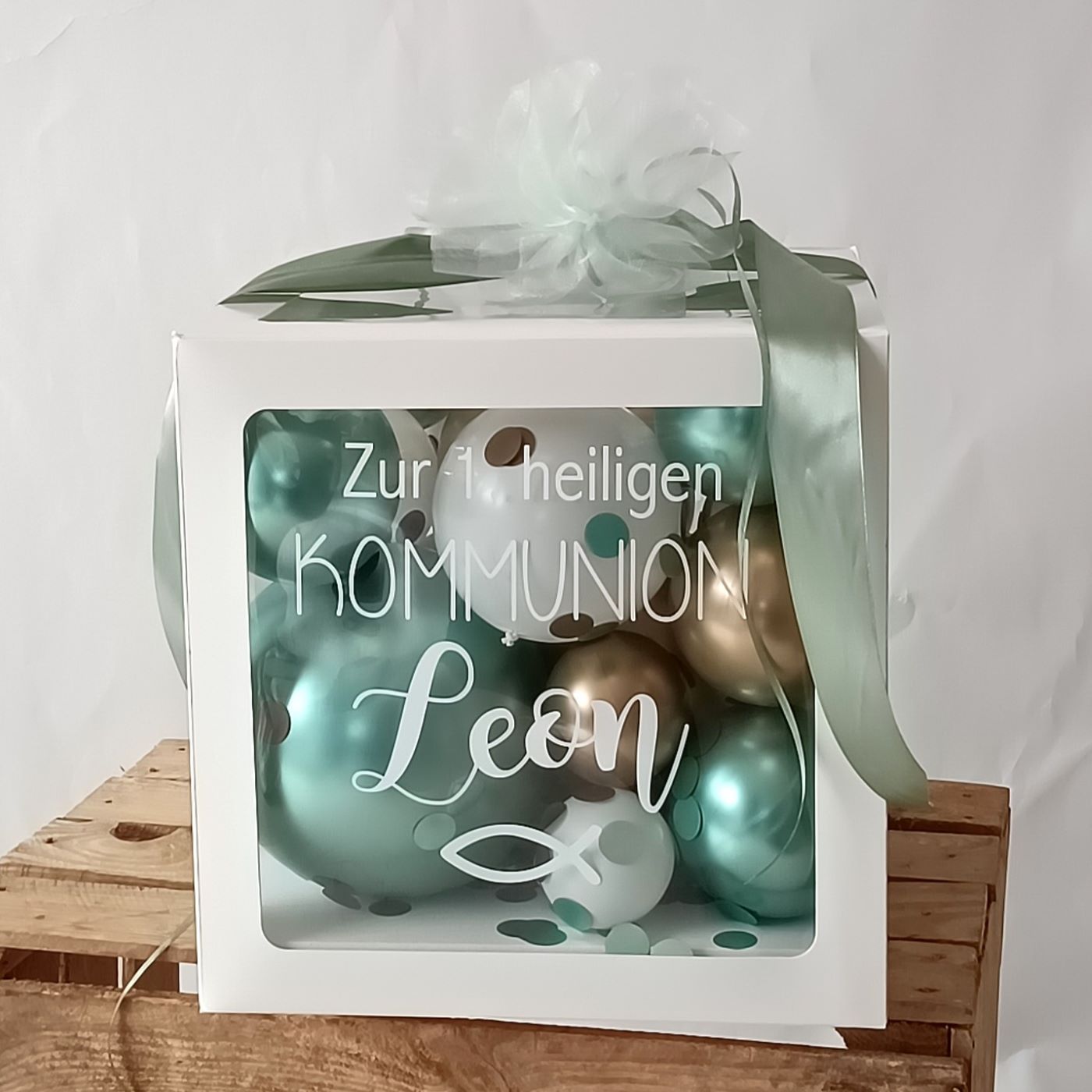 Quadratische Geschenkbox gefüllt mit metallicfarbenen Ballons in Blau, Mint und Gold, vorne Aufdruck 'Zur 1. heiligen Kommunion Leon' mit Fischsymbol
