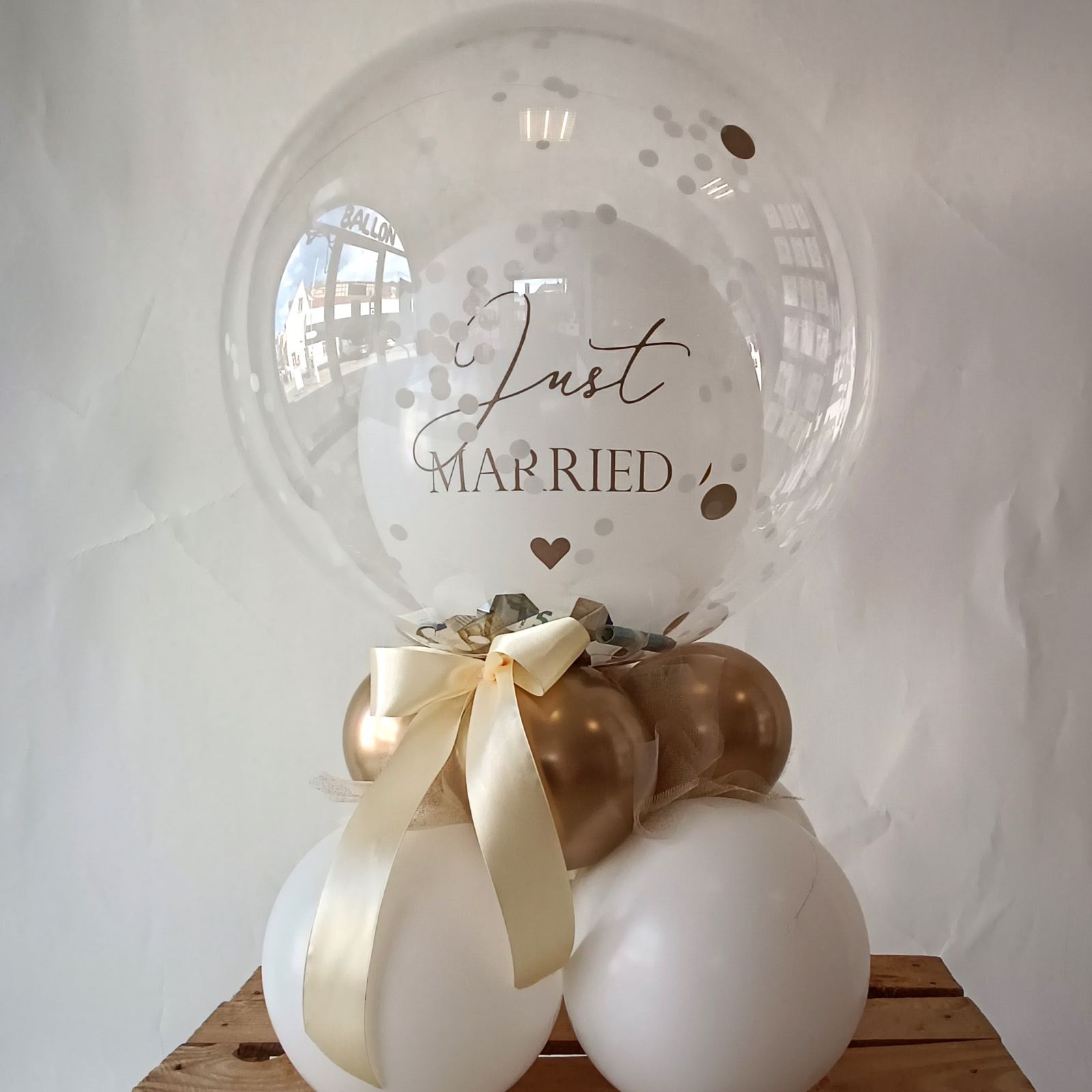 Transparenter Bubbleballon mit 'Just Married'-Aufdruck, dekoriert mit weißen und goldenen Ballons sowie Schleife