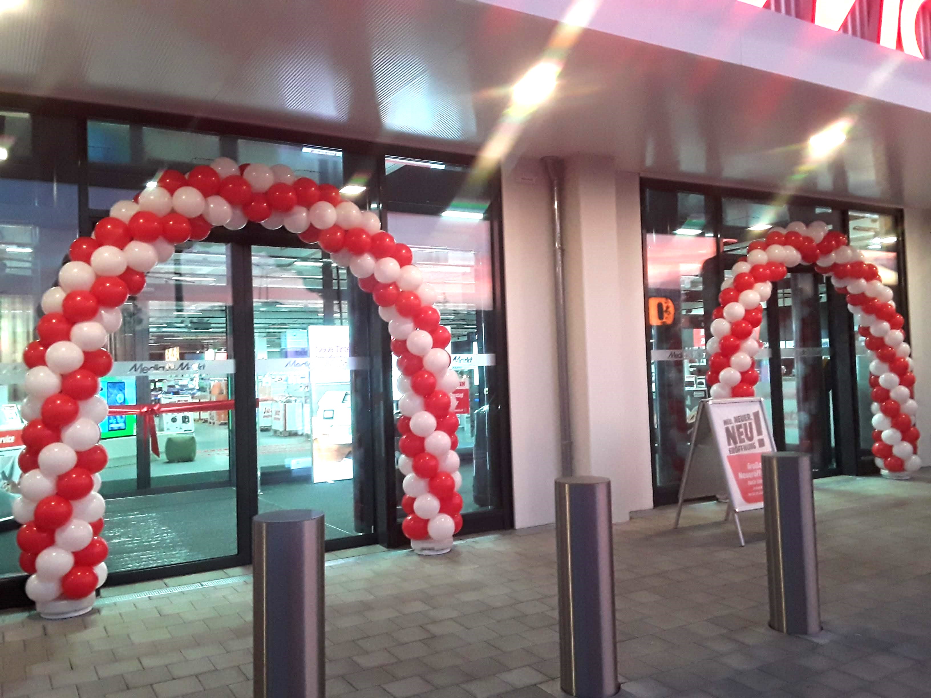 2 gedrehte Ballonbögen jeweils in Rot und Weiß über einem automatischen Eingang