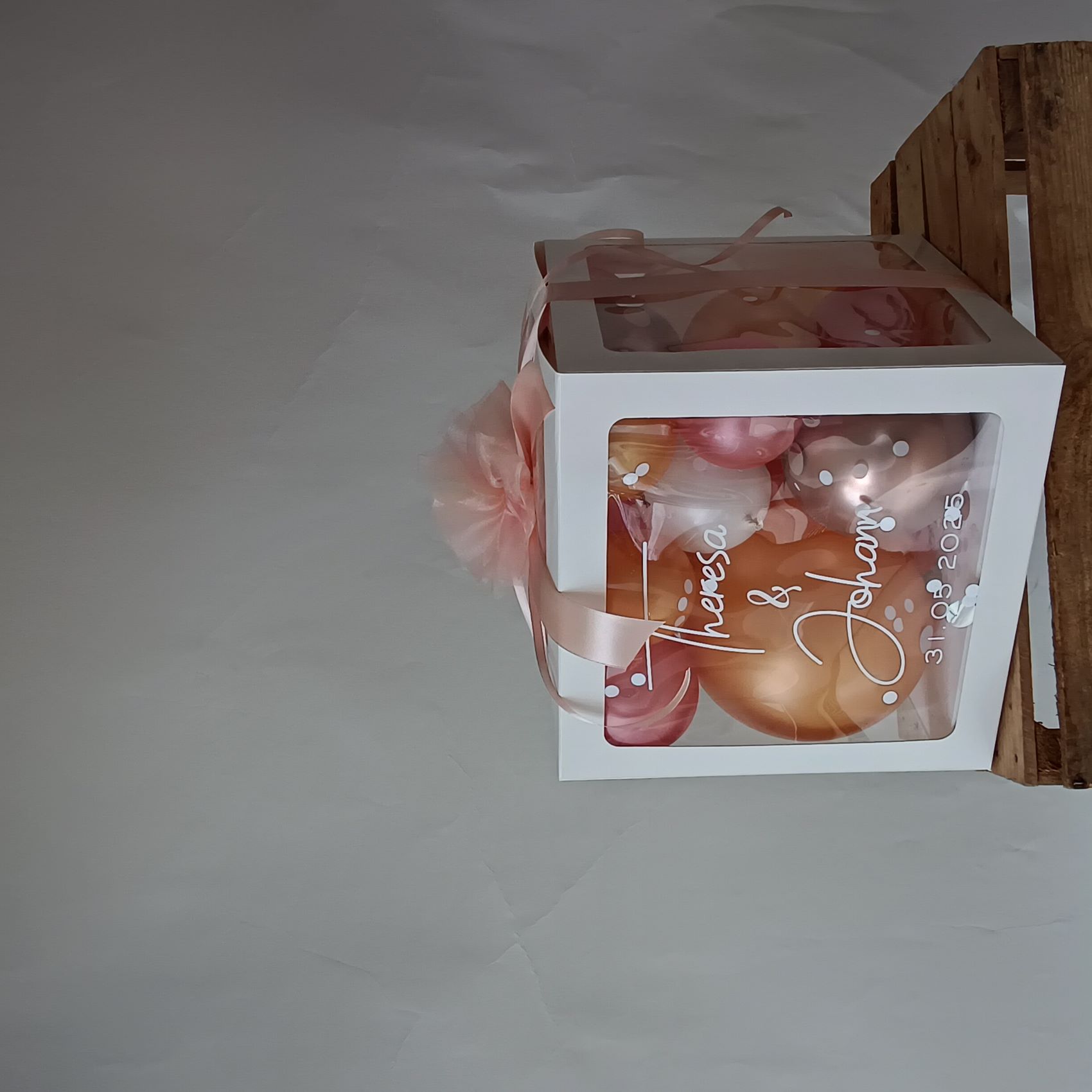 Ballonbox mit roséfarbenen, weißen und apricotfarbenen Ballons, Schleife und Tüllblume, Aufdruck mit Namen und Hochzeitsdatum.