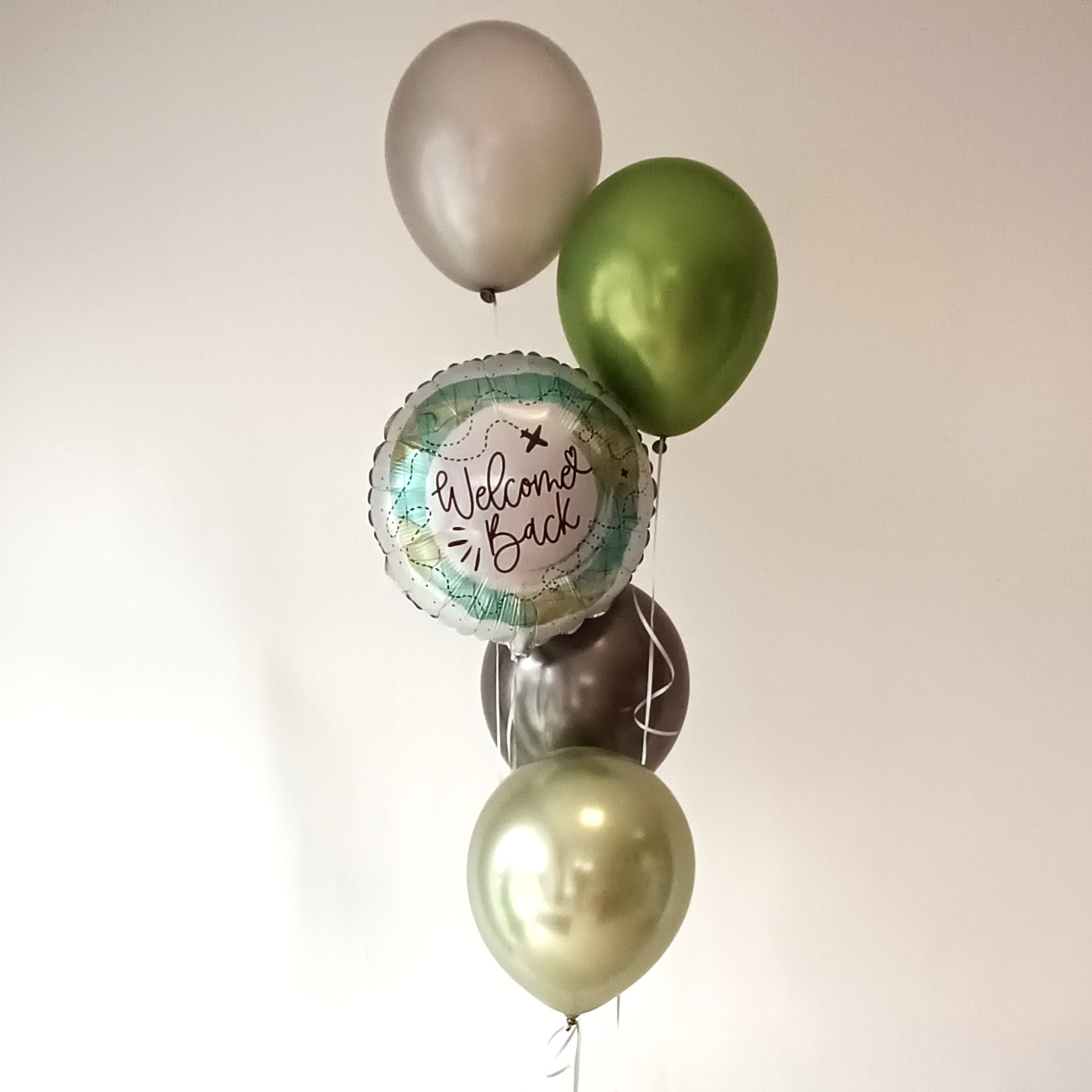 Ballonstrauß mit fünf Luftballons in Grün, Silber, Schwarz und einem runden Folienballon mit „Welcome Back“-Aufdruck, arrangiert in Helium-Bouquet-Form