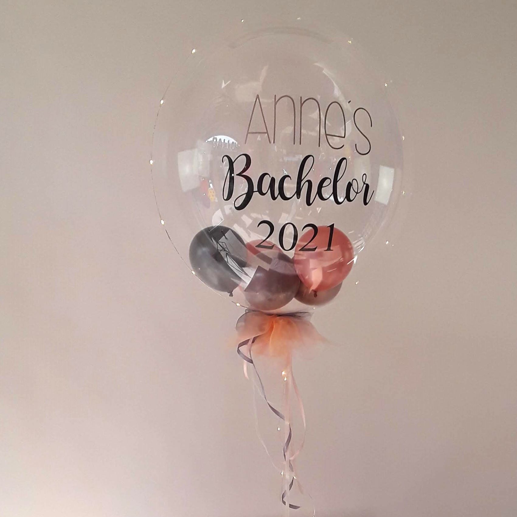 Klarer Bubble-Ballon mit schwarzem Aufdruck „Anne’s Bachelor 2021“, innen Mini-Ballons in Roségold und Anthrazit, unten Tüllschleife; um die Kugel verläuft eine feine LED-Lichterkette.