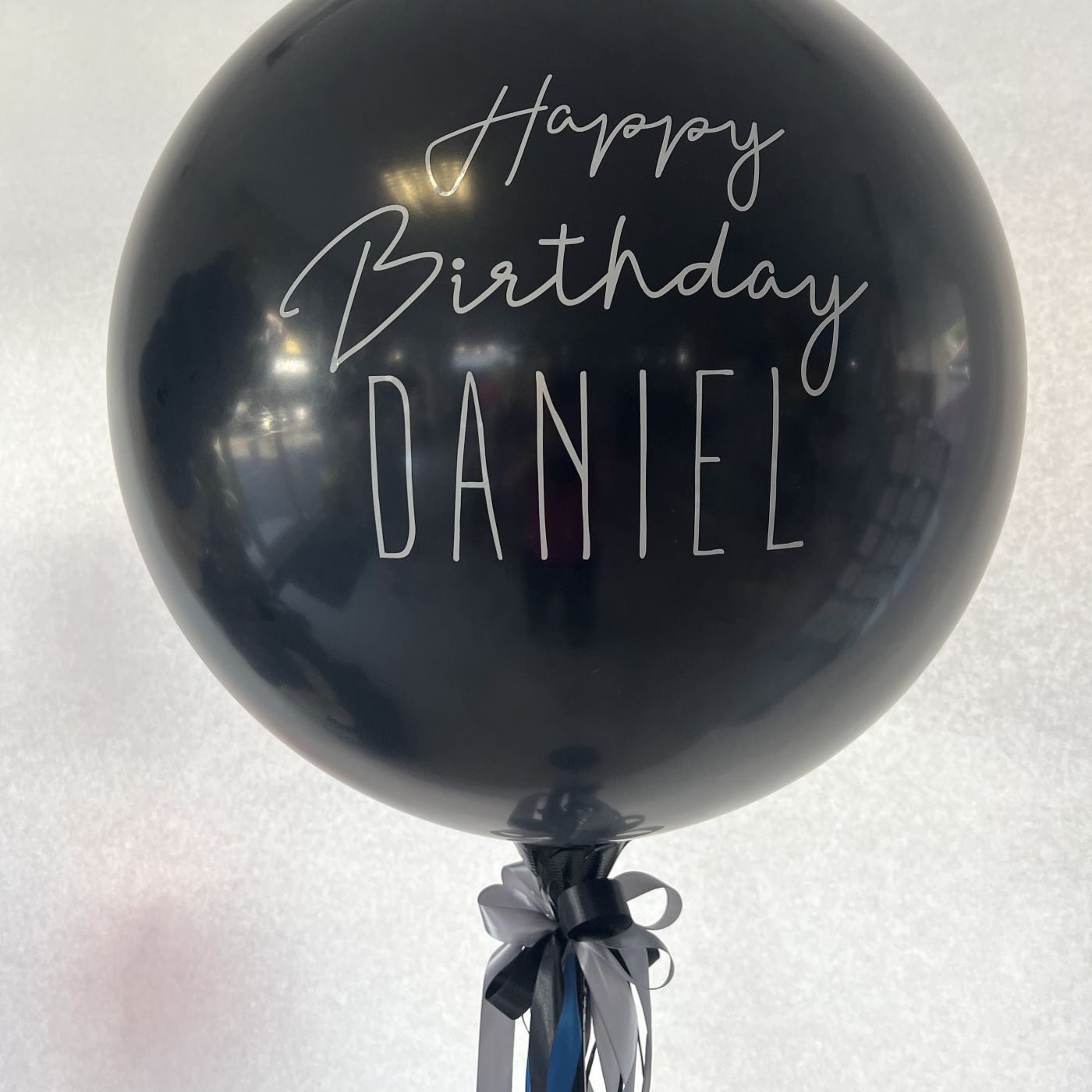 Große schwarze Ballonkugel mit weißem Aufdruck 'Happy Birthday Daniel' und dekorativer Schleife aus Satinbändern in Schwarz und Blau.