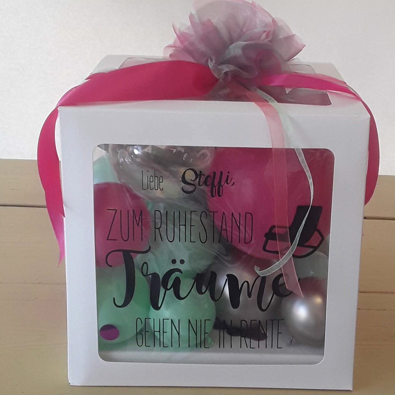 Weiße Geschenkbox mit Sichtfenster, gefüllt mit bunten Ballons und Federn, Aufdruck 'Liebe Steffi, zum Ruhestand Träume gehen nie in Rente'.