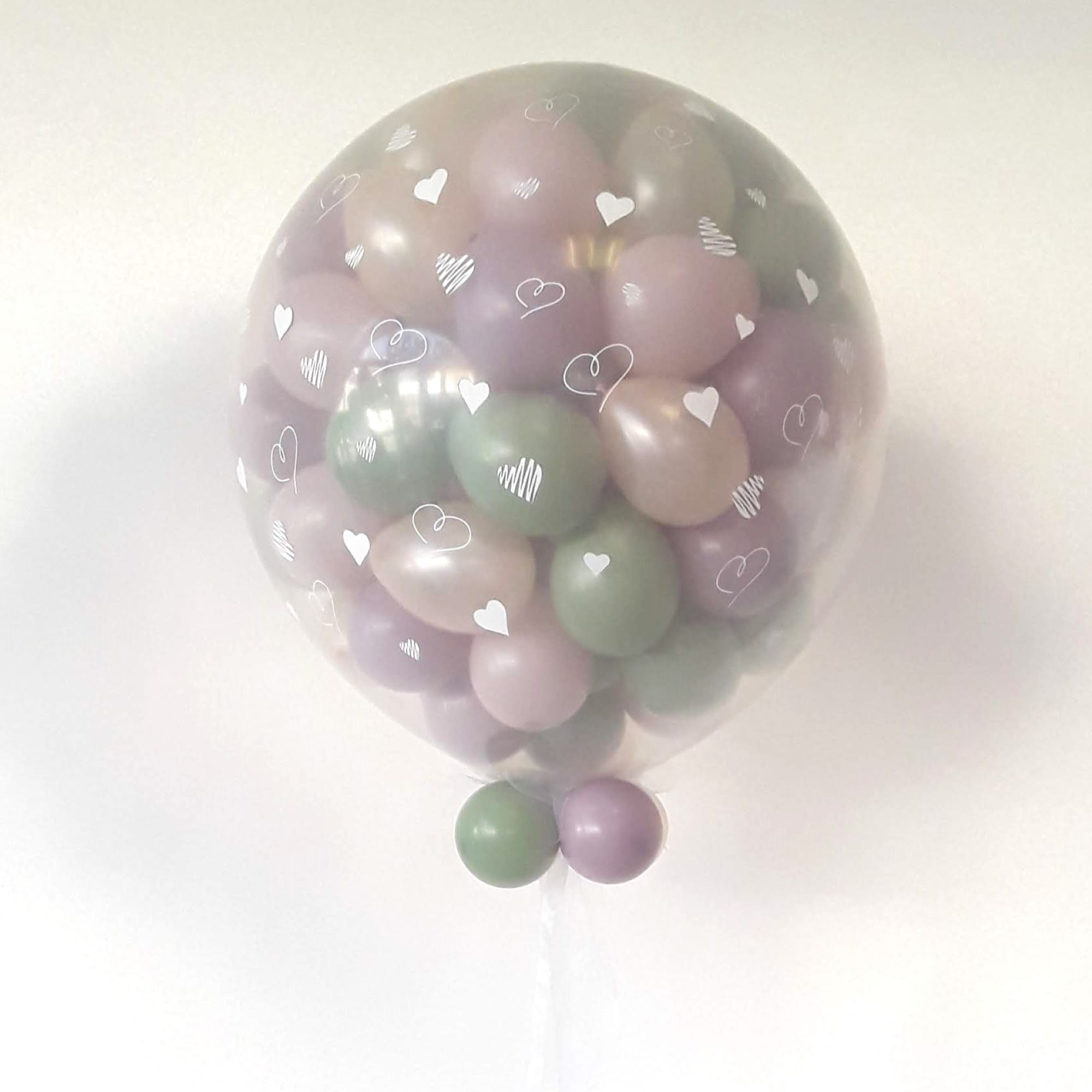 Nahaufnahme eines transparenten ca. 80‑cm‑Ballons mit weißen Herzmotiven, gefüllt mit ca. 30 kleinen Pastellballons in Lila, Rosé und Salbeigrün; unten zwei kleine Ballons in Grün und Lila als Abschluss, weißer Hintergrund