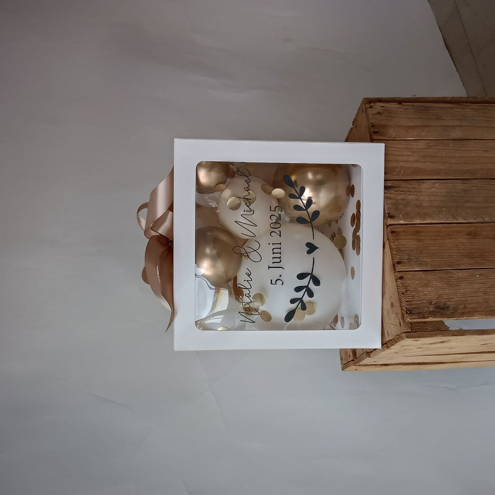 Ballonbox mit goldenen und cremefarbenen Luftballons, goldener Schleife, schwarzem Blätteraufdruck und Konfetti.