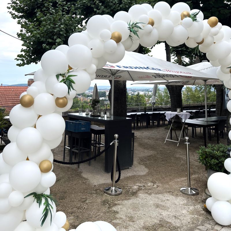 Eleganter, komplett weißer Ballonbogen mit goldenen Akzentballons und grünen Blättern, aufgebaut vor einem verglasten Gebäude2