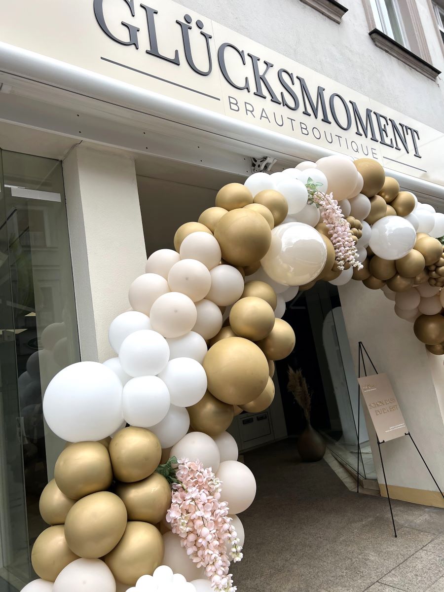 Ballonbogen in Weiß und Gold mit floralem Akzent vor dem Shop "Glücksmoment"
