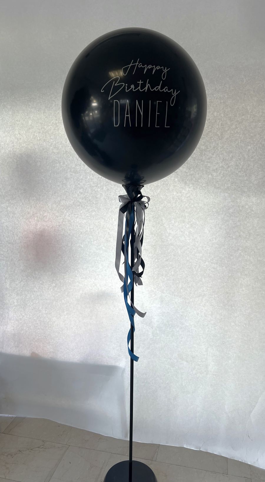 Große schwarze Ballonkugel mit weißem Aufdruck 'Happy Birthday Daniel' und dekorativer Schleife aus Satinbändern in Schwarz und Blau und eine Schleife