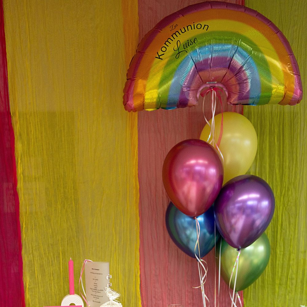 Regenbogen-Folienballon mit Aufdruck 'Zur Kommunion Luise' an einem Band mit bunten Latexballons, bunt gedeckter Tisch in den Farben rot, gelb, grün auf weißer Tischdecke2