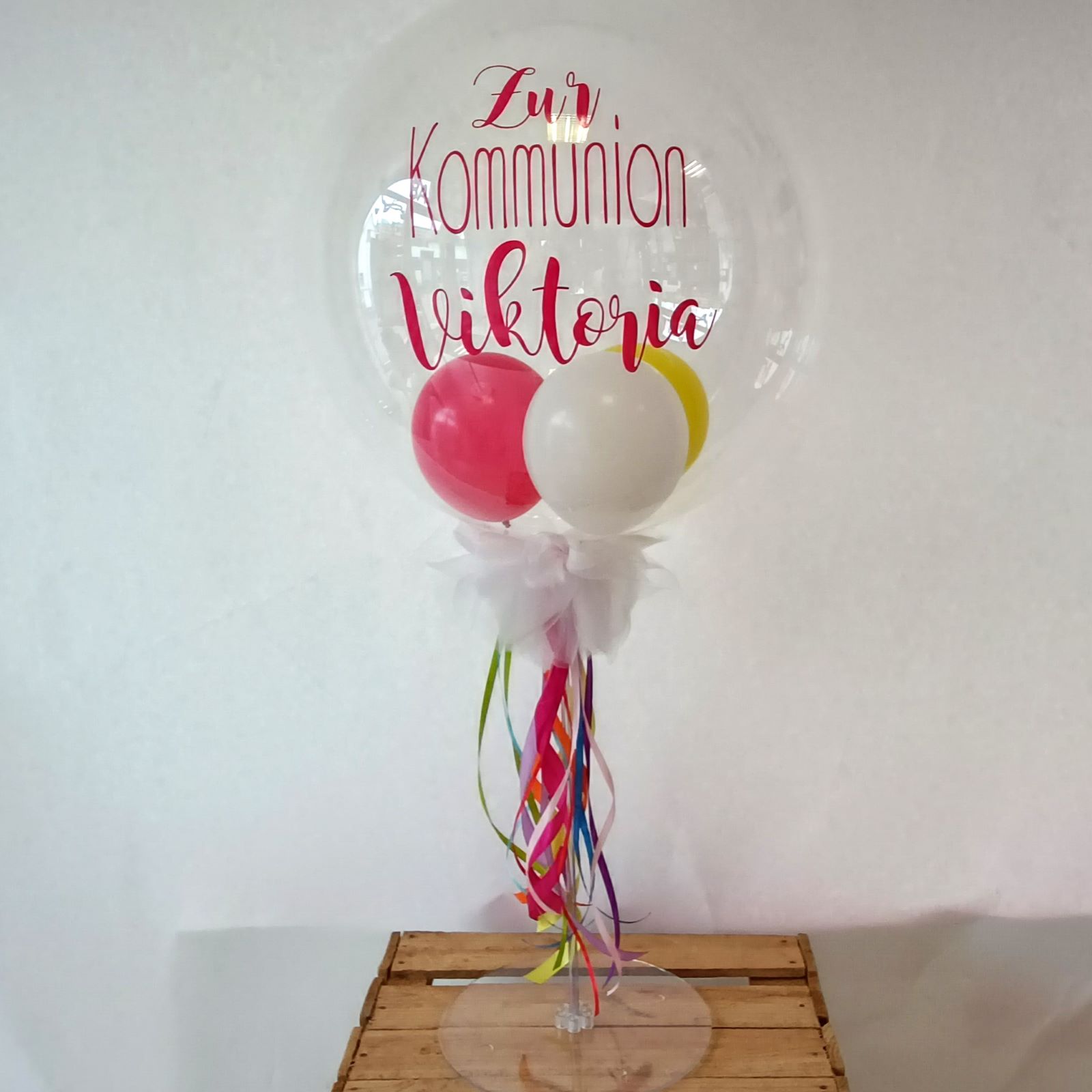 Bubble-Ballon mit Aufdruck 'Zur Kommunion Viktoria' gefüllt mit Latexballons in den Farben pink, weiß, gelb, Tüll in weiß und bunte Schleifenbänder auf Tischständer