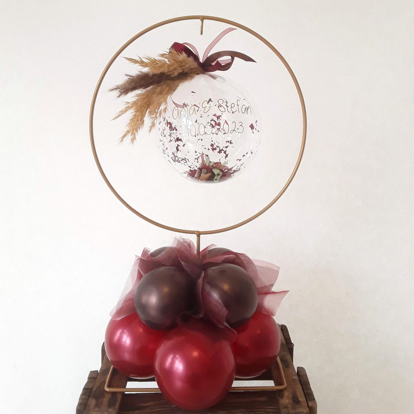 Kleiner transparenter Ballon gefüllt mit Geldscheinen udn Konfetti mit Aufdruck 'Anja & Stefan 10.05.2023' im Ringgestell  mit Trockenblumen und Federn, Ballonbasis aus Aubergine- und magentafarbenen Mini-Ballons und Tüll