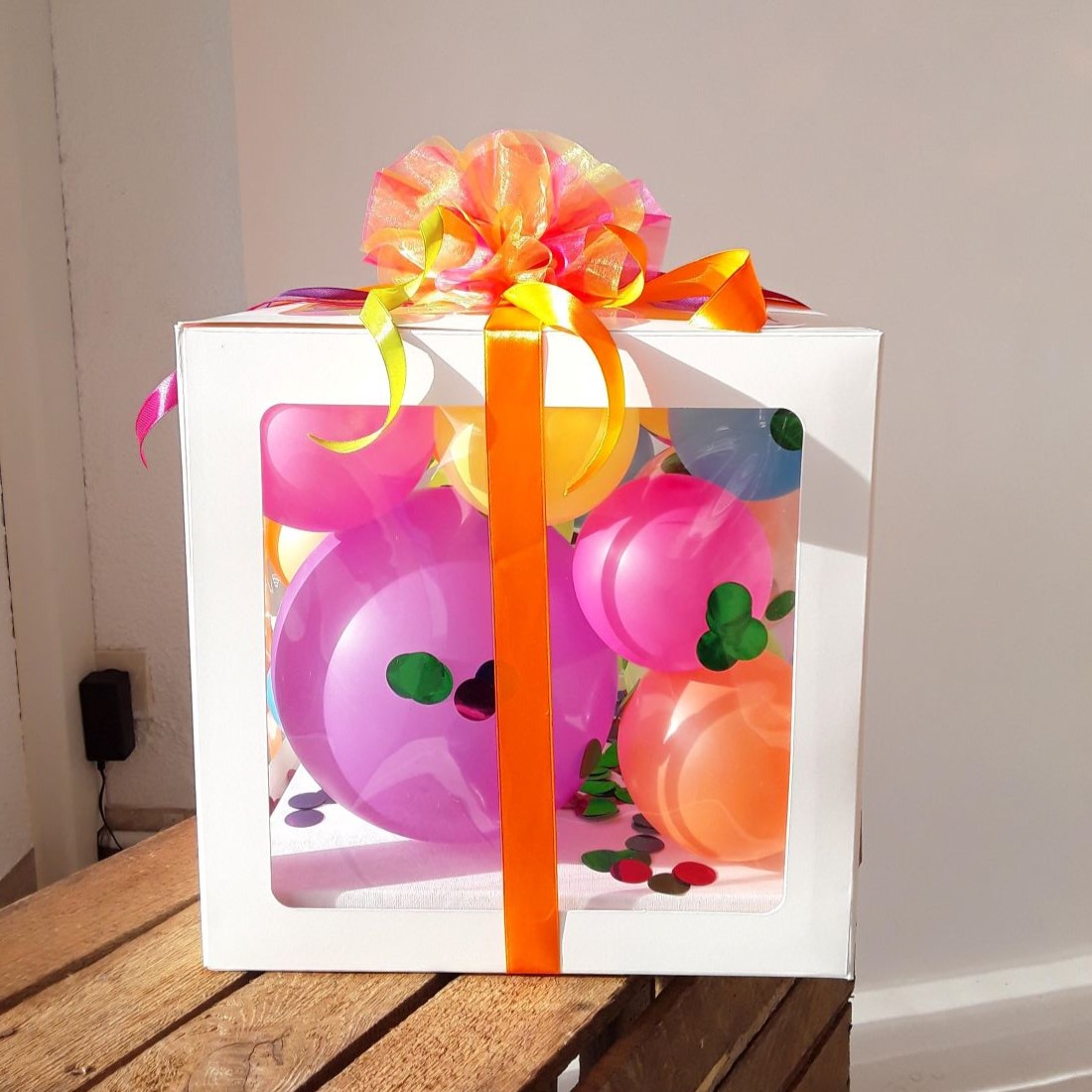 Weiße Ballonbox mit Ballons in Lila, Orange, Gelb, Pink und Grün, dekoriert mit bunter Schleife und Konfetti.