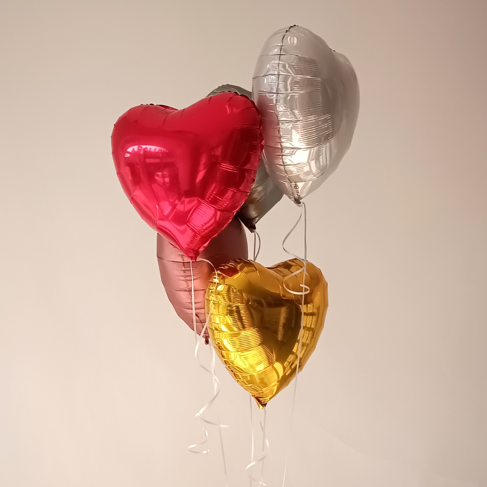 Mehrere Herz-Folienballons in Metallic-Farben (Gold, Silber, Rot, Rosa), mit weißen Bändern, vor neutralem Hintergrund2