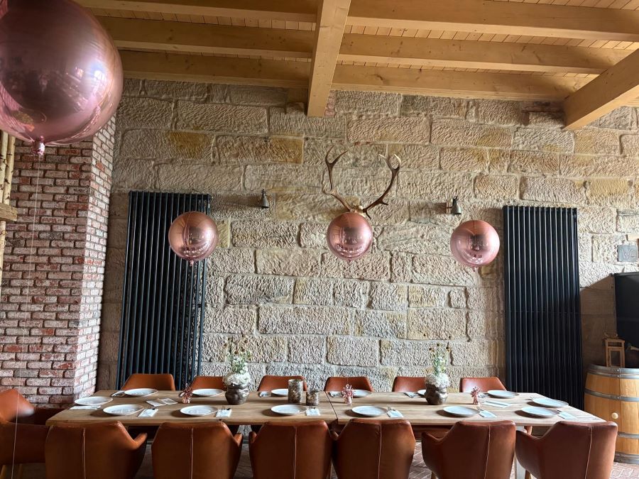 4 Schwebende Orb-Ballons in glänzendem Roségold als elegante Tischdekoration über einer festlich gedeckten Tafel in einem Raum mit Mauer- und Holzwänden