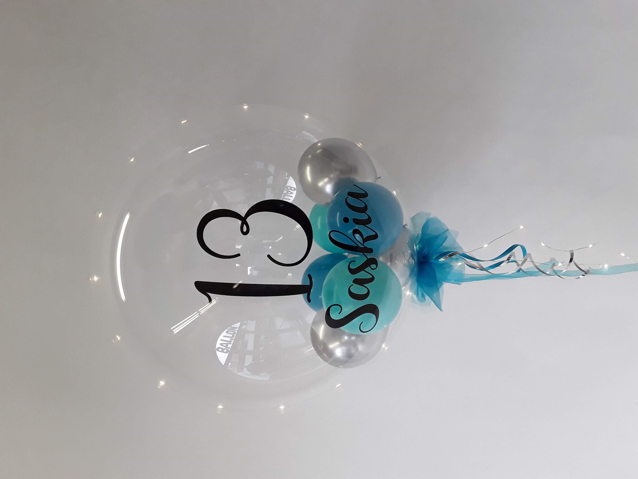 Transparenter Bubble-Ballon mit schwarzer Aufschrift „13 Saskia“, gefüllt mit silbernen und türkisfarbenen Ballons, dekoriert mit blauer Tüllschleife, silbernem Band und integrierter Lichterkette.