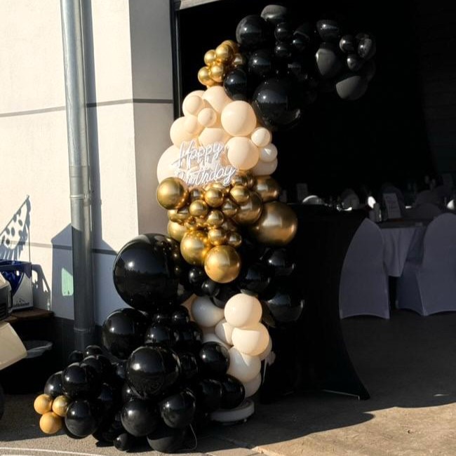 Halbkreisbogen aus schwarzen, goldenen und champagnerfarbenen Ballons in verschiedenen Größen, mittig ein leuchtender Neon-Schriftzug „Happy Birthday“, aufgebaut vor einem Gebäude im Freien