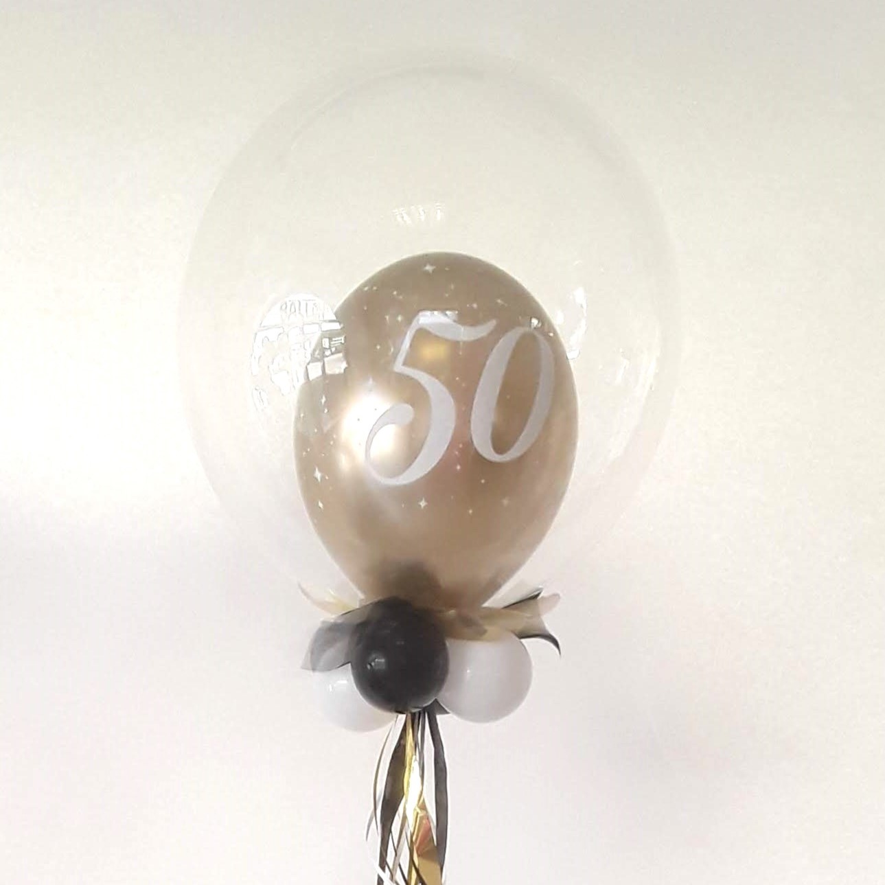 Transparenter Bubble-Ballon mit goldenem Innenballon und großer weißer Zahl „50“, dekoriert mit kleinen weißen Sternchen, schwarzen und weißen Mini-Ballons sowie schwarzem und goldenem Geschenkband.