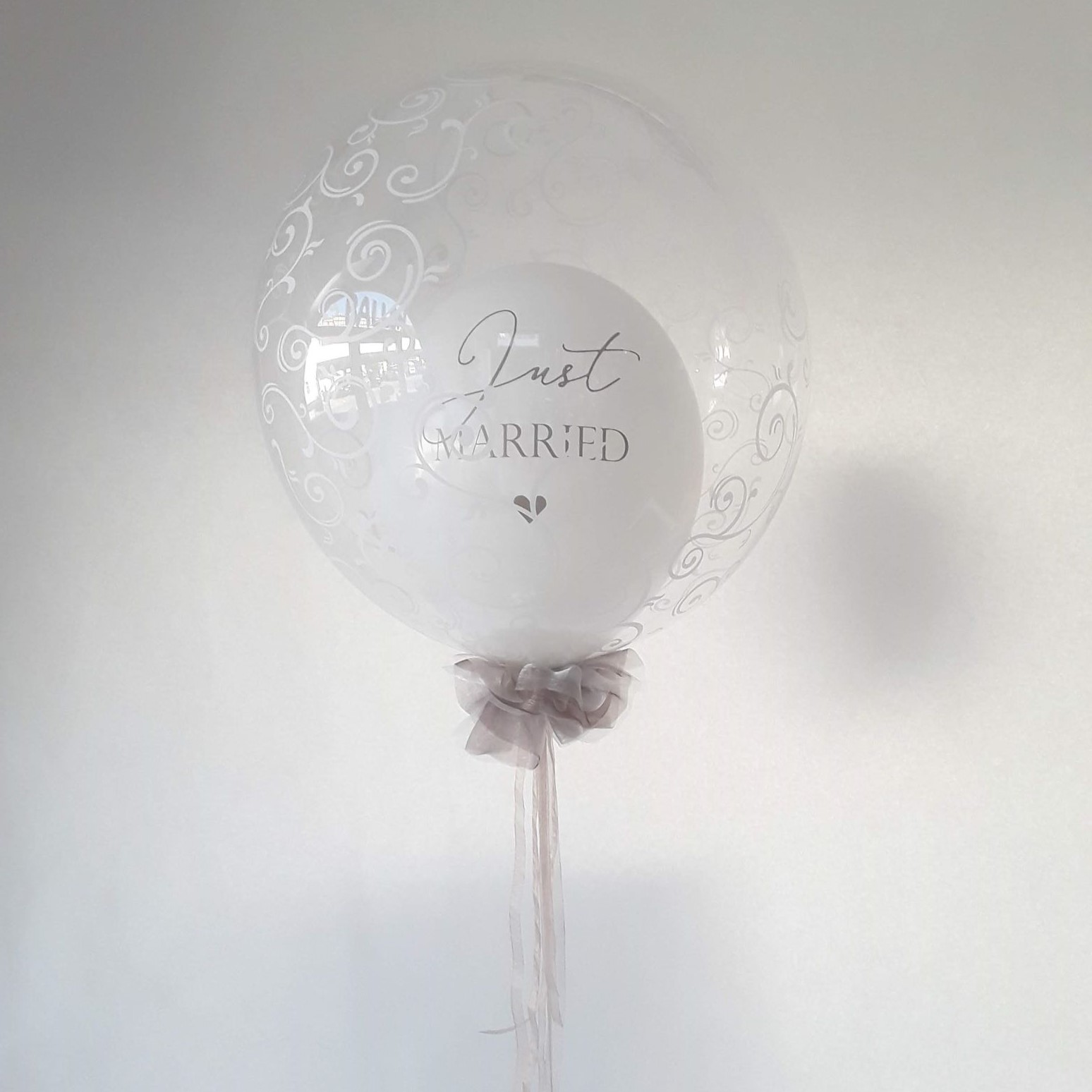 Transparenter Bubble-Ballon mit weißem Innenballon und der Aufschrift „Just Married“, verziert mit weißen Ornamenten und einer eleganten Schleife aus Tüll.
