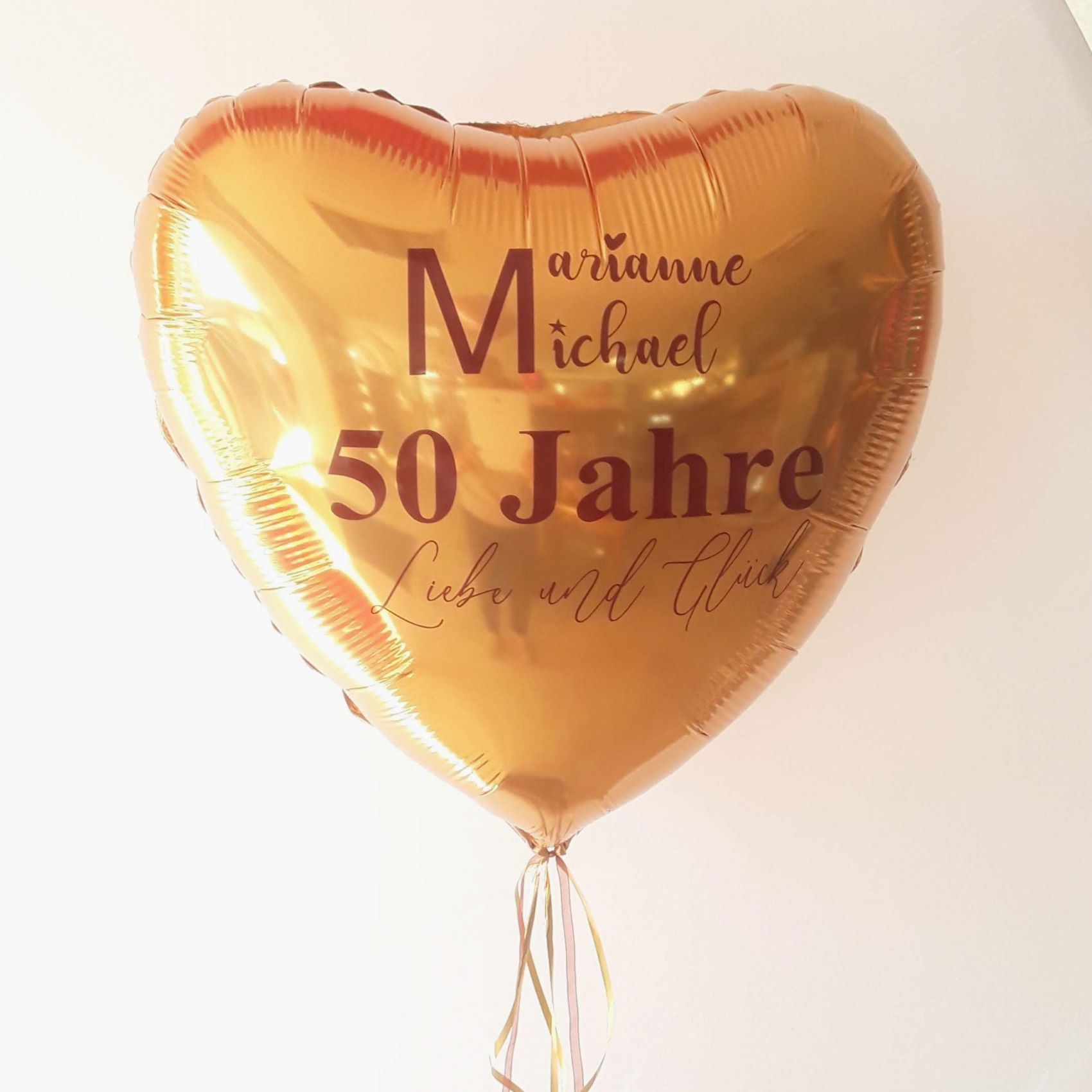 Goldener Herz-Folienballon mit roter Schrift '50 Jahre Liebe und Glück' sowie Namen, mit schmalen goldenen Bändern.