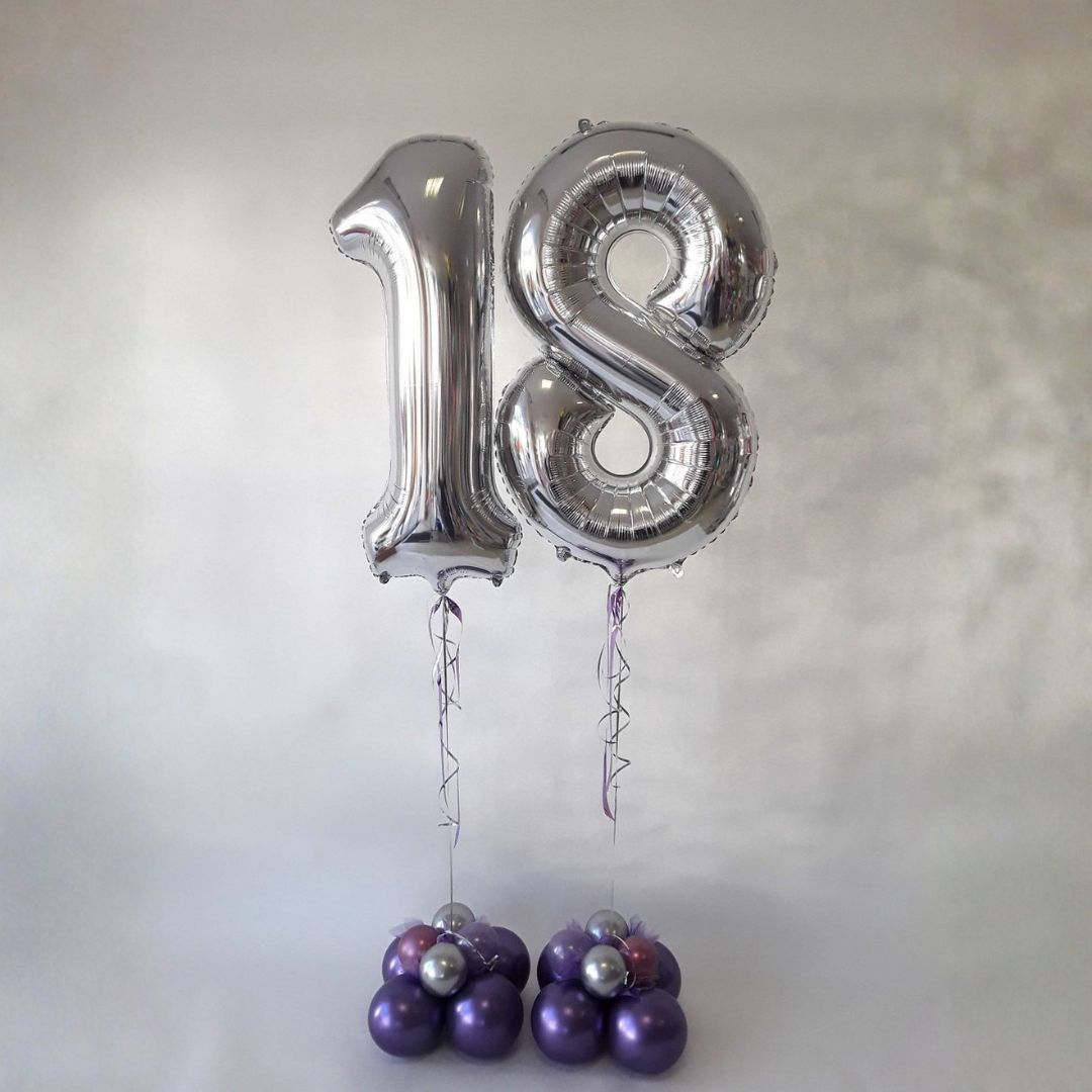 Silberne Folienballons in Form der Zahlen 1 und 8 mit violett-silbernem Ballongewicht auf hellem Hintergrund.