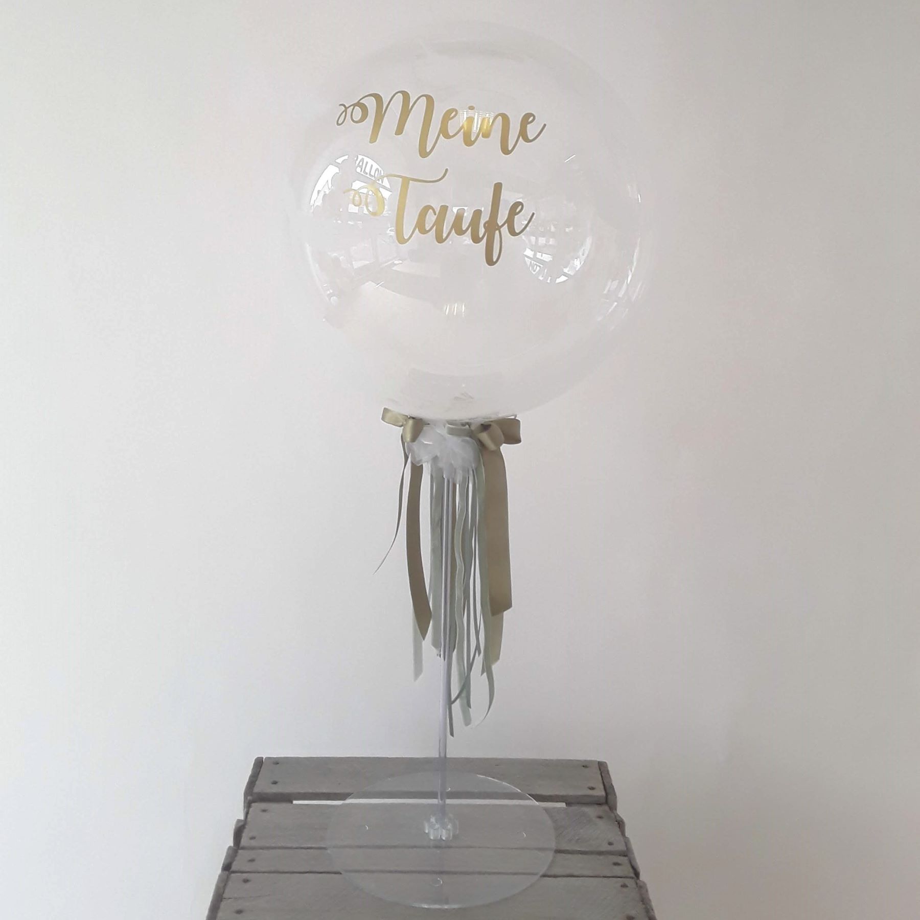 Transparenter Bubbleballon mit goldenem Aufdruck 'Meine Taufe', dekoriert mit weißen und goldenen Bändern, auf Ständer.