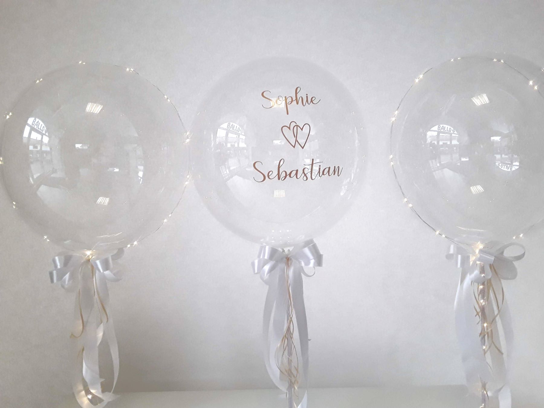 Transparenter Hochzeitsballon mit Namen 'Sophie & Sebastian', Tischständer mit Schleife in Weiß und Lichterkette, zwei unbedruckte, transparente Ballon-Tischständer