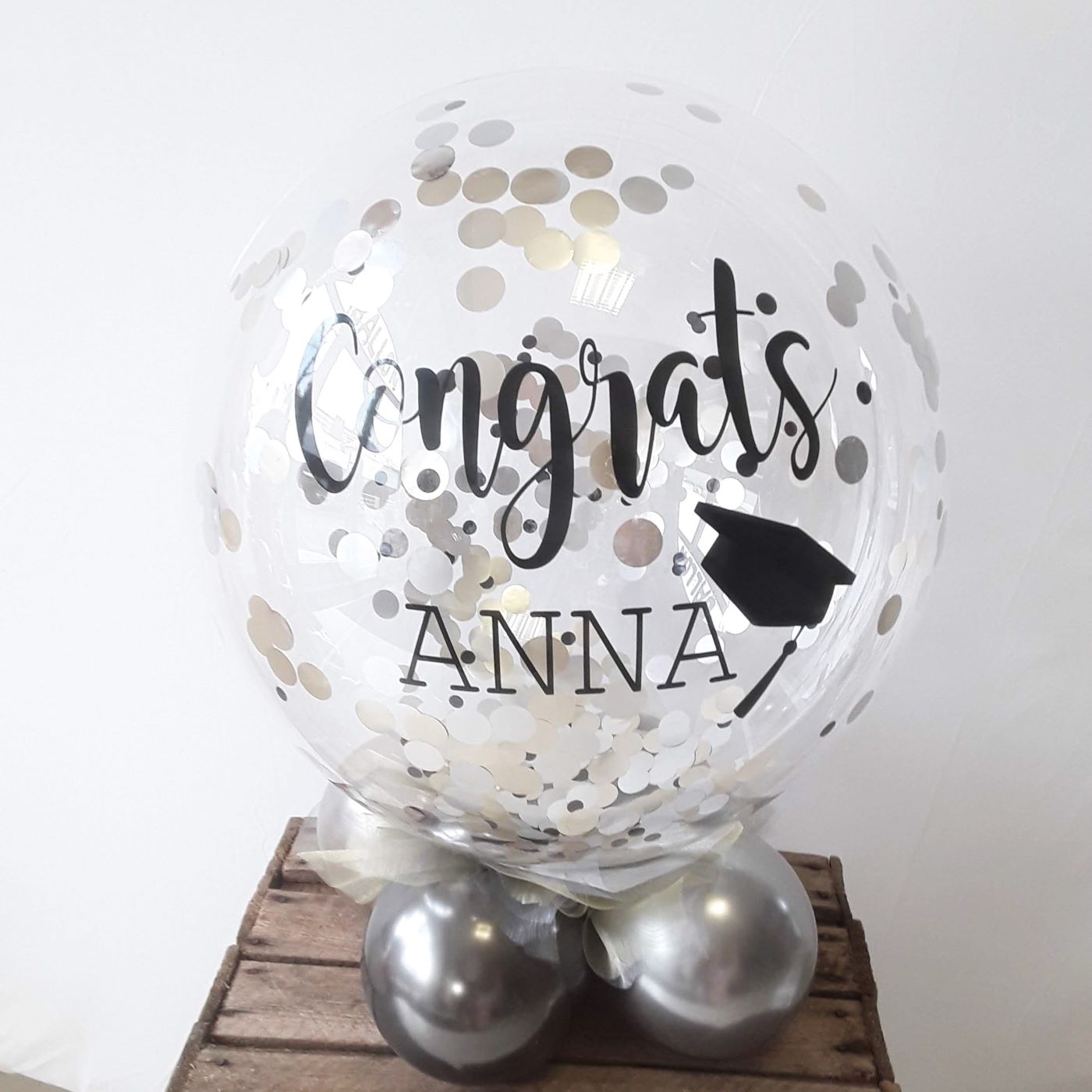 Konfetti-Bubble mit schwarzem Schriftzug „Congrats Anna“ und kleinem Graduierten-Hut, luftgefüllt auf einem silbernen Ballonfuß als Tischbouquet.