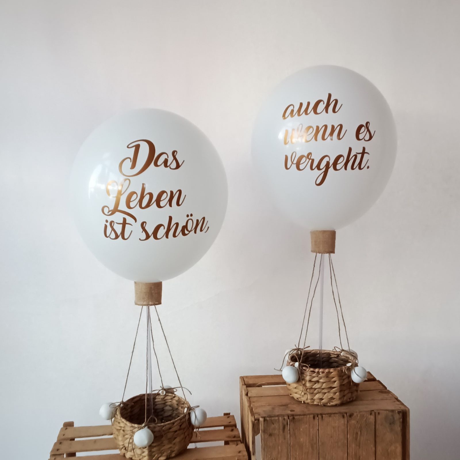 Zwei weiße Ballons in Heißluftballon-Optik mit goldener Aufschrift 'Das Leben ist schön' und 'auch wenn es vergeht', befestigt an kleinen Körben.