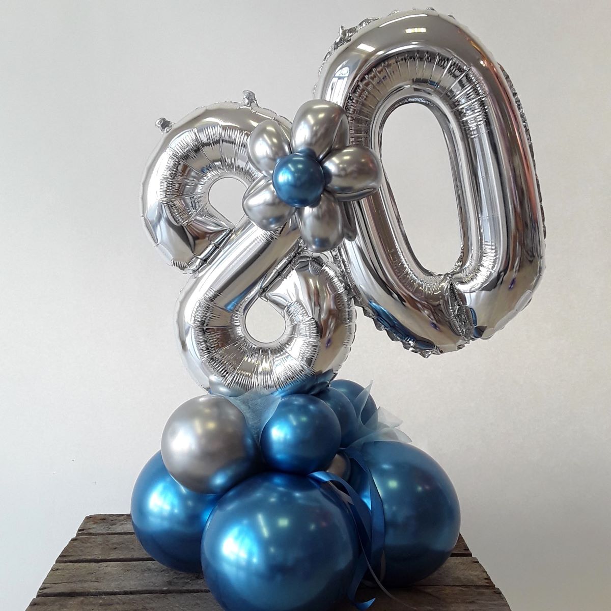 Ballon-Arrangement mit großen silbernen Zahlenballons '8' und '0' auf einer Basis aus glänzenden blauen Ballons, dekoriert mit einer silber-blauen Ballonblume.