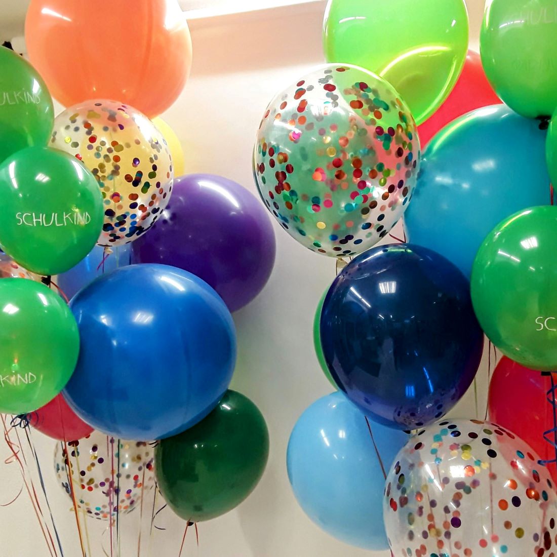 Zwei bunte Ballonbouquets mit Aufdruck 'Schulkind' und Konfettiballons, auf weißem Hintergrund fotografiert.