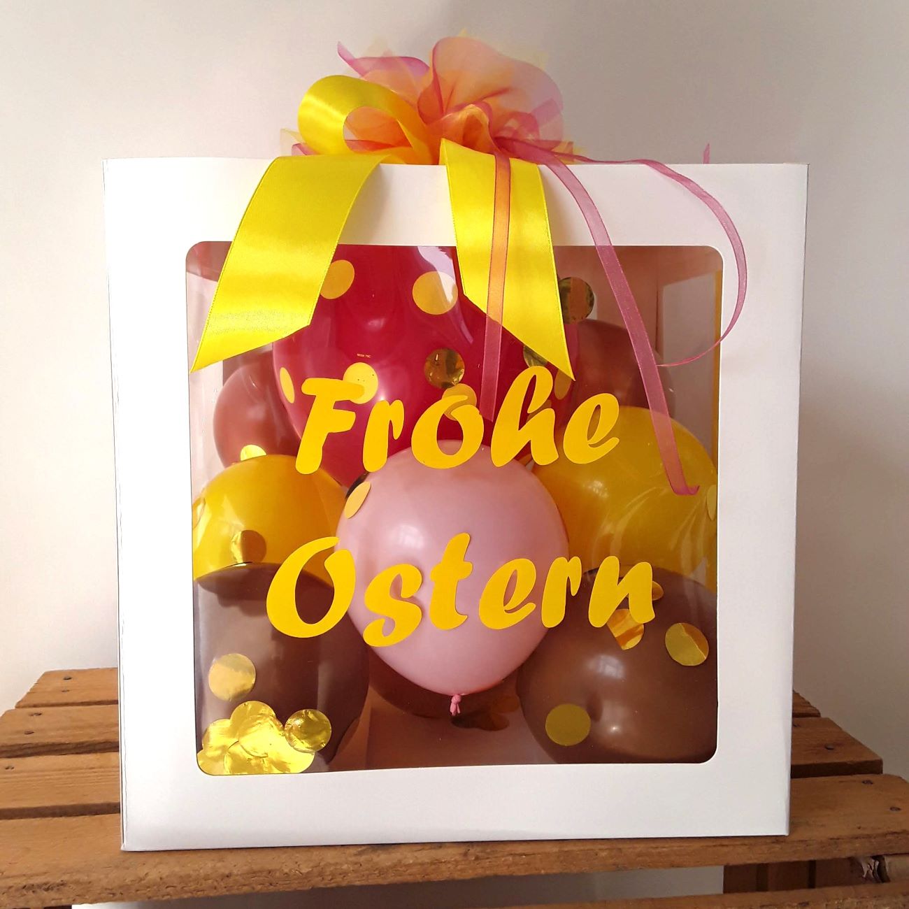 Ballonbox aus weißem Karton mit transparenten Fenstern, gelbem Aufdruck "Frohe Ostern", kleinen Ballons in pink, rosa, gelb und braun, goldenem Konfetti und gelb-pinker Schleife