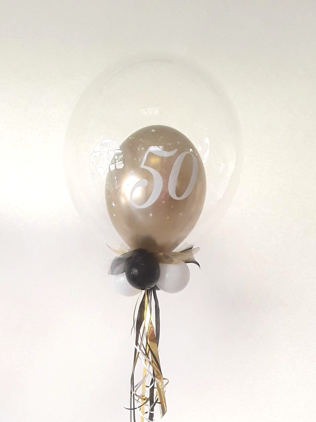 Transparenter Bubble-Ballon mit goldenem Innenballon und großer weißer Zahl „50“, dekoriert mit kleinen weißen Sternchen, schwarzen und weißen Mini-Ballons sowie schwarzem und goldenem Geschenkband.