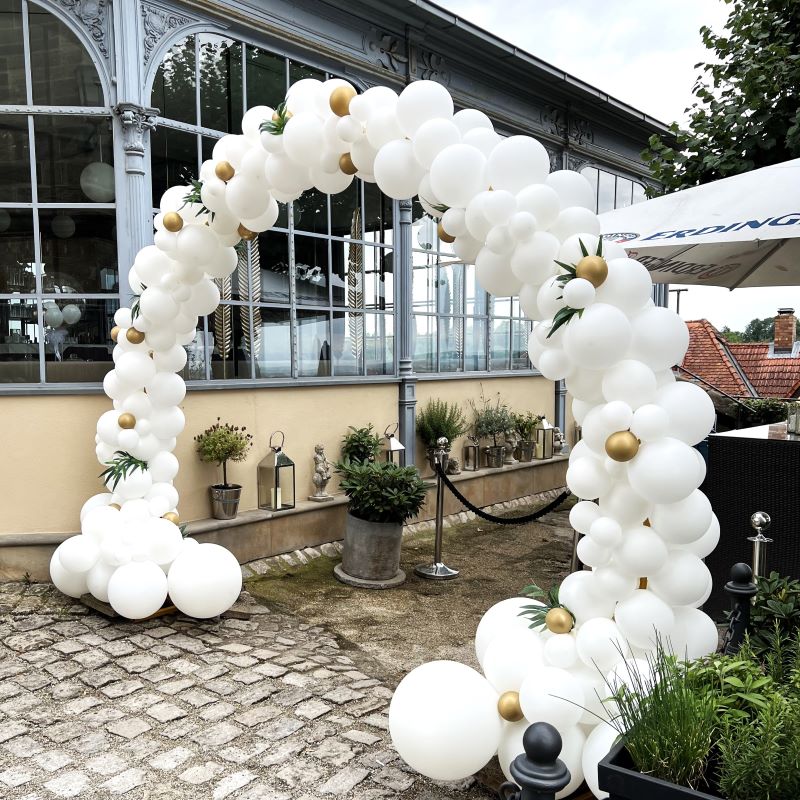 Eleganter, komplett weißer Ballonbogen mit goldenen Akzentballons und grünen Blättern, aufgebaut vor einem verglasten Gebäude3