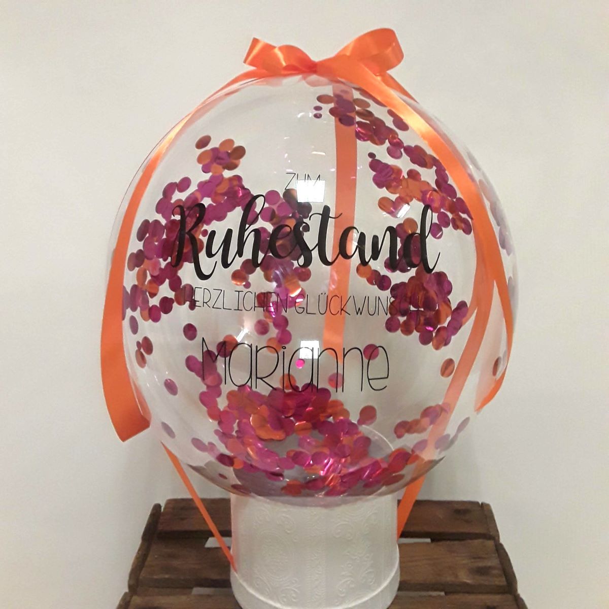 Bubble-Ballon mit pink-orangem Konfetti, orangefarbenem Schleifenband und Aufdruck 'Zum Ruhestand herzlichen Glückwunsch Marianne'.