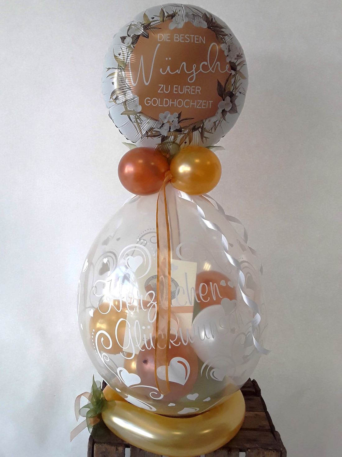Runder Folienballon mit Goldhochzeitsmotiv über einem transparenten Bubbleballon mit weißen Herzmustern und goldenen Innenballons, dekoriert mit Bändern und goldenen Akzenten.